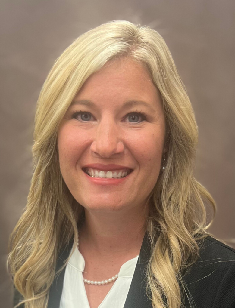 A woman with long blonde hair, wearing a black blazer, a white blouse, and a pearl necklace, smiles at the camera against a plain background.